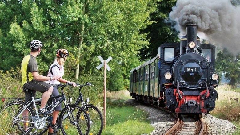 Two cyclists wait at a level crossing while a steam locomotive passes by.