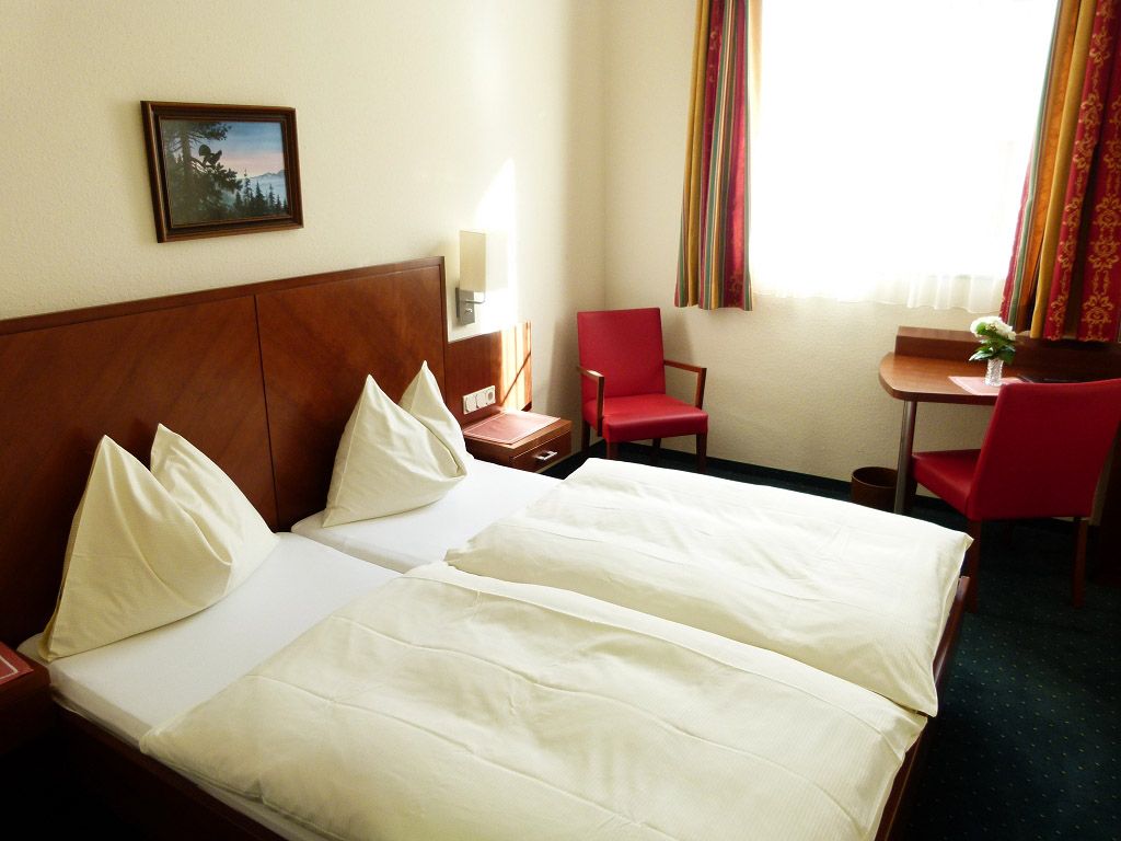 A hotel room with two single beds, red chairs and a small table by the window.