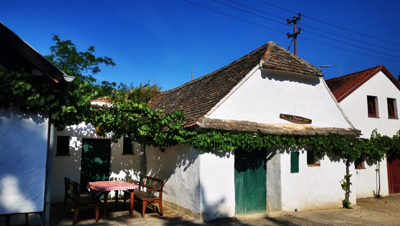 White building in a wine cellar lane with vines and outdoor table.
