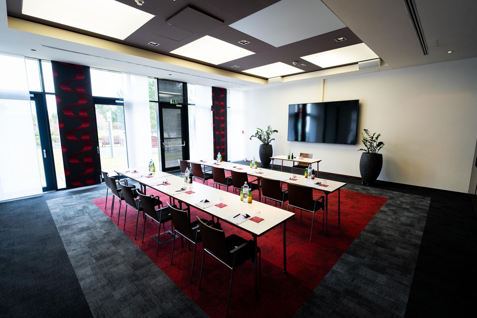 A modern conference room with tables, chairs, and a large screen on the wall. The room has large windows and plants as decoration.