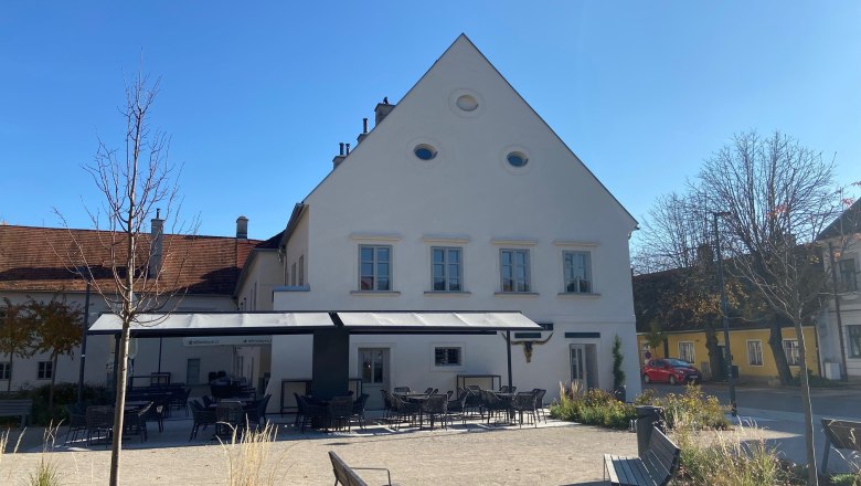 A garden with empty tables and chairs in front of a white building with a pointed roof on a sunny day.