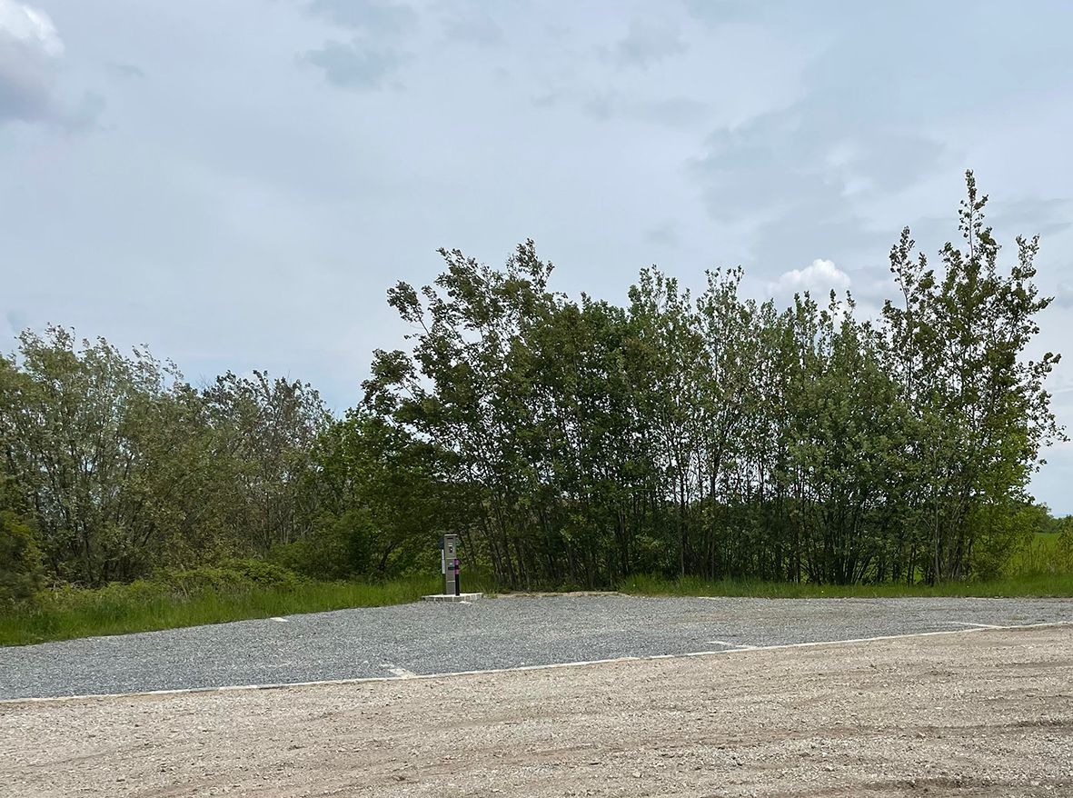 An empty motorhome pitch with a gravel floor, surrounded by trees and a cloudy sky.