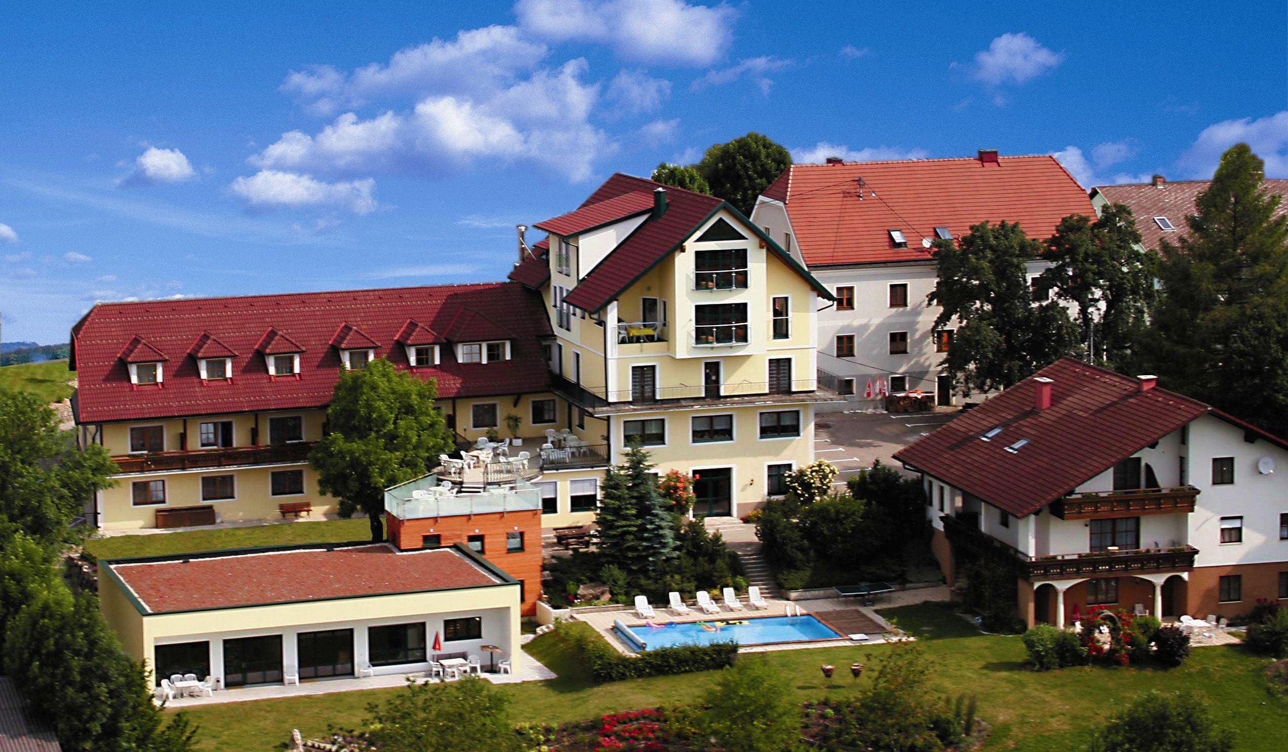 A hotel complex with red roofs, pool and garden, surrounded by trees and flowering plants under a blue sky.