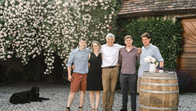 A family stands smiling in front of a flowering wall, with a dog in the background and a wine barrel with flowers on the right.