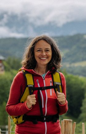 Irene Beckmann steht vor einer malerischen Hügelkulisse mit einem traditionellen Gebäude im Hintergrund. Nebelschwaden und bewaldete Berge vervollständigen die idyllische Szene, die Freude an der Natur und kultureller Entdeckung vermittelt.