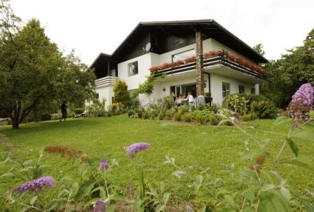 A two-storey white house with a balcony, surrounded by a green garden with flowers and trees.