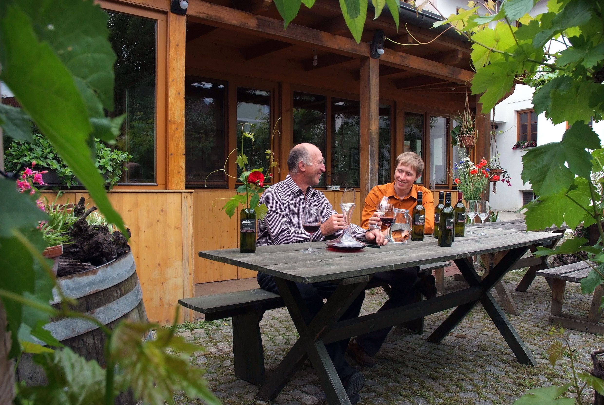 Two men sit at a wooden table outside and taste wine. Surrounded by plants and a wooden building in the background.