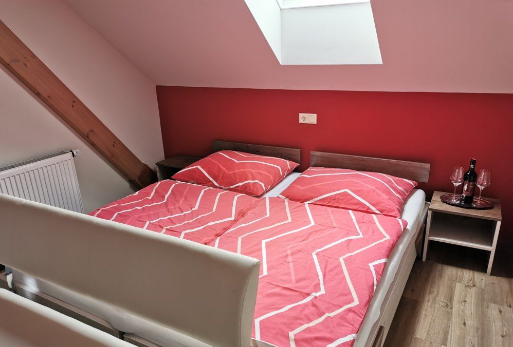 Bedroom with red wall, double bed with red bed linen, skylight and bedside table with wine bottle and glasses.