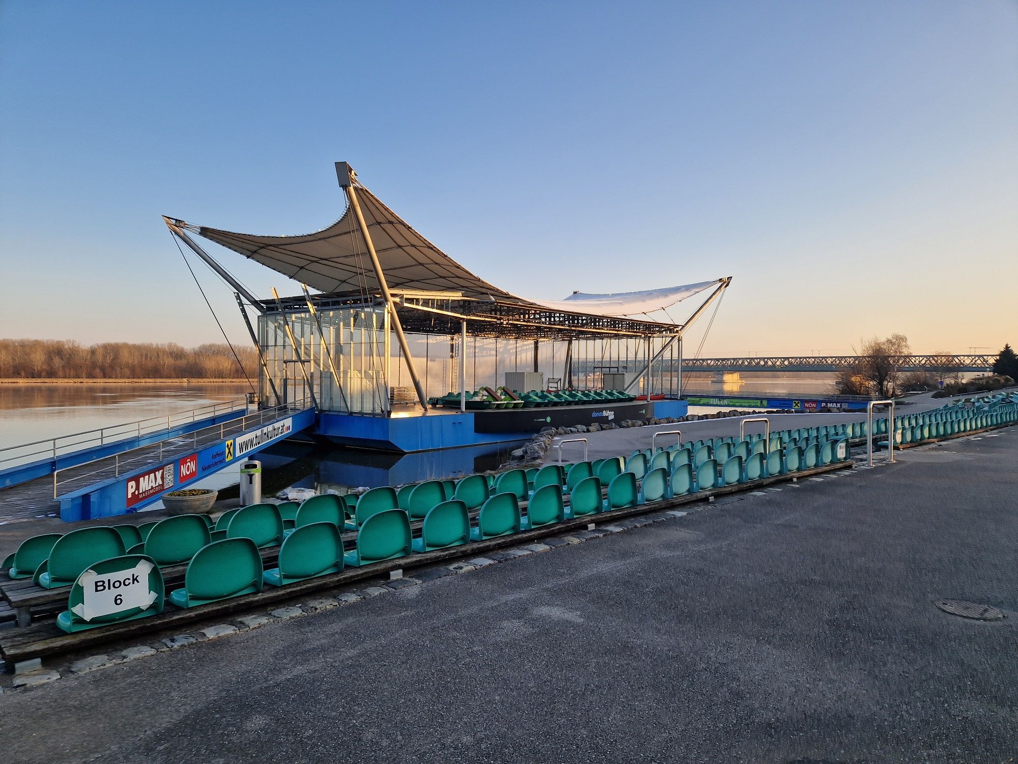 The Donaubühne Tulln with empty rows of seats and a modern roof, with the Danube and a bridge in the background.