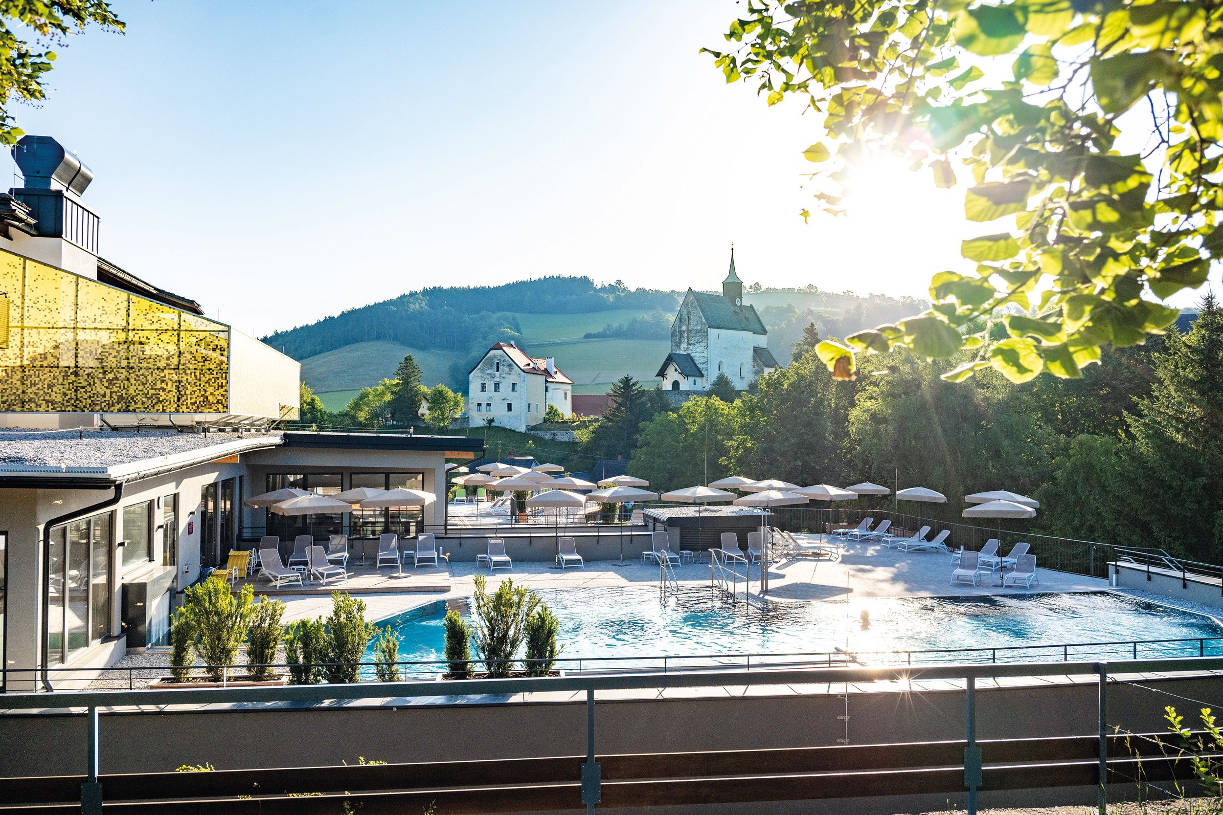Outdoor area of a hotel with pool, parasols and sun loungers, surrounded by nature and a church in the background.