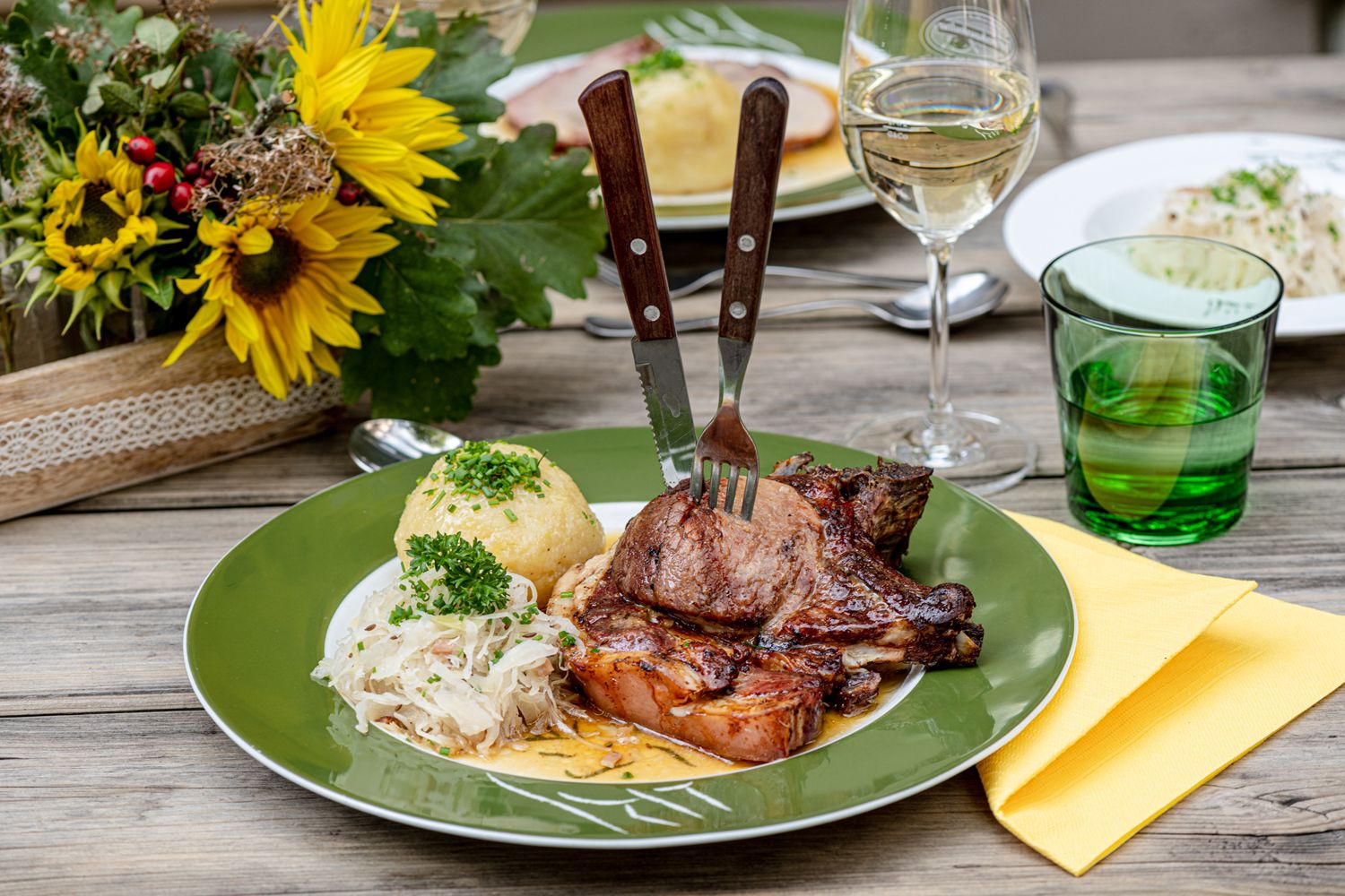 A plate of roast pork, dumplings and sauerkraut on a wooden table, next to it a glass of white wine and a flower arrangement.