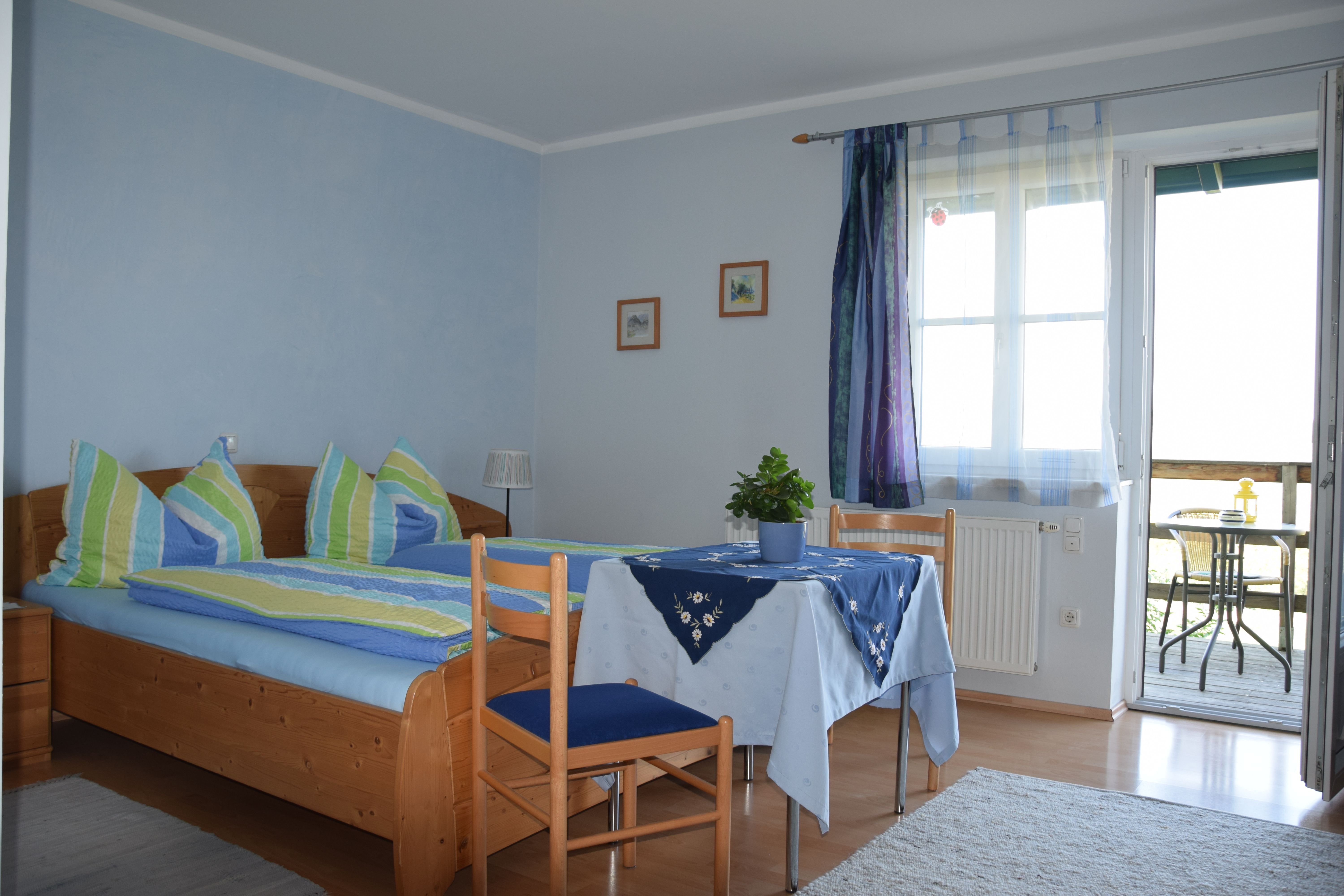 Cozy guest room with double bed, table and chairs, blue curtains and access to a balcony with seating.