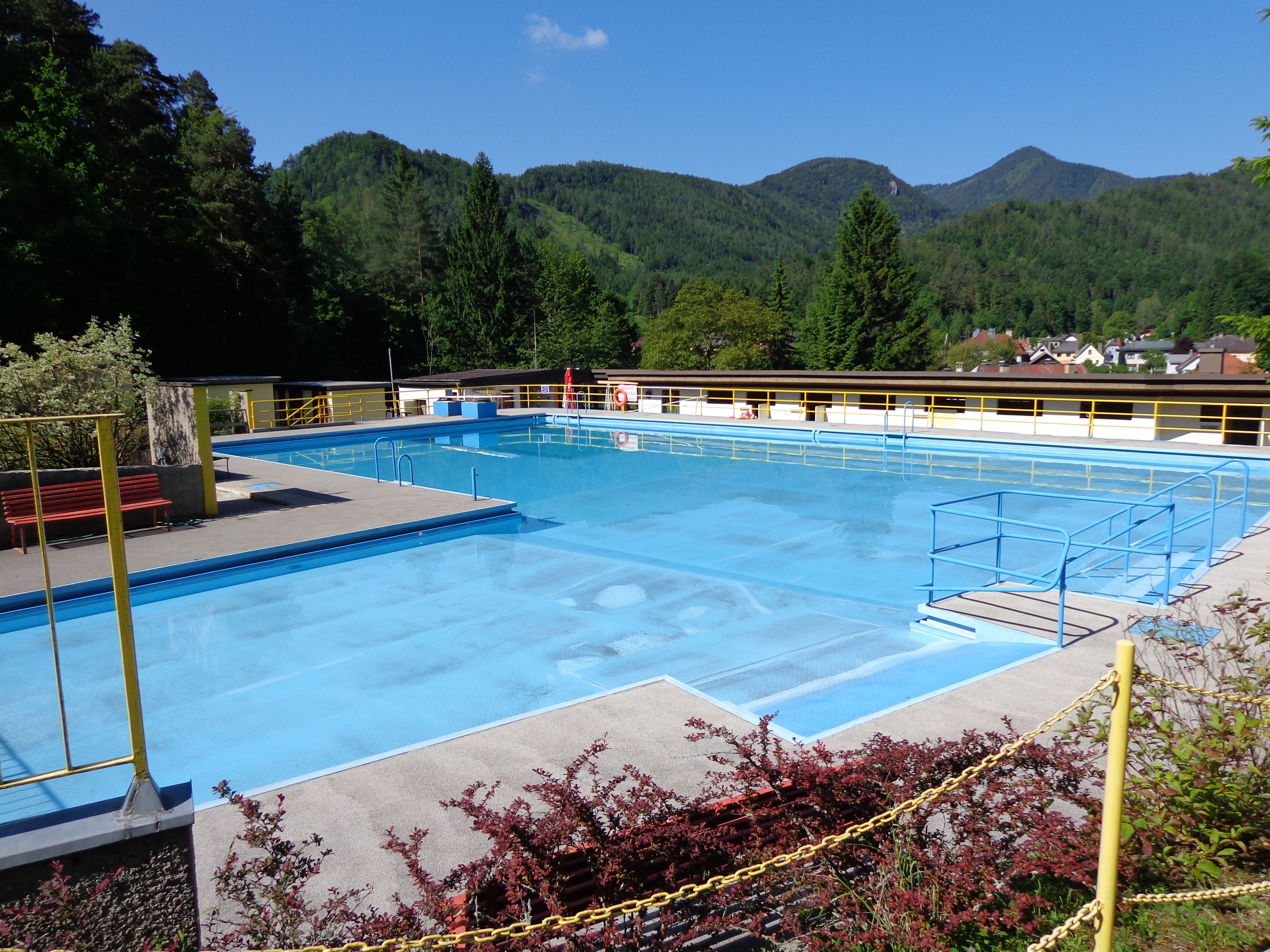 An empty outdoor pool with blue water, surrounded by mountains and trees.