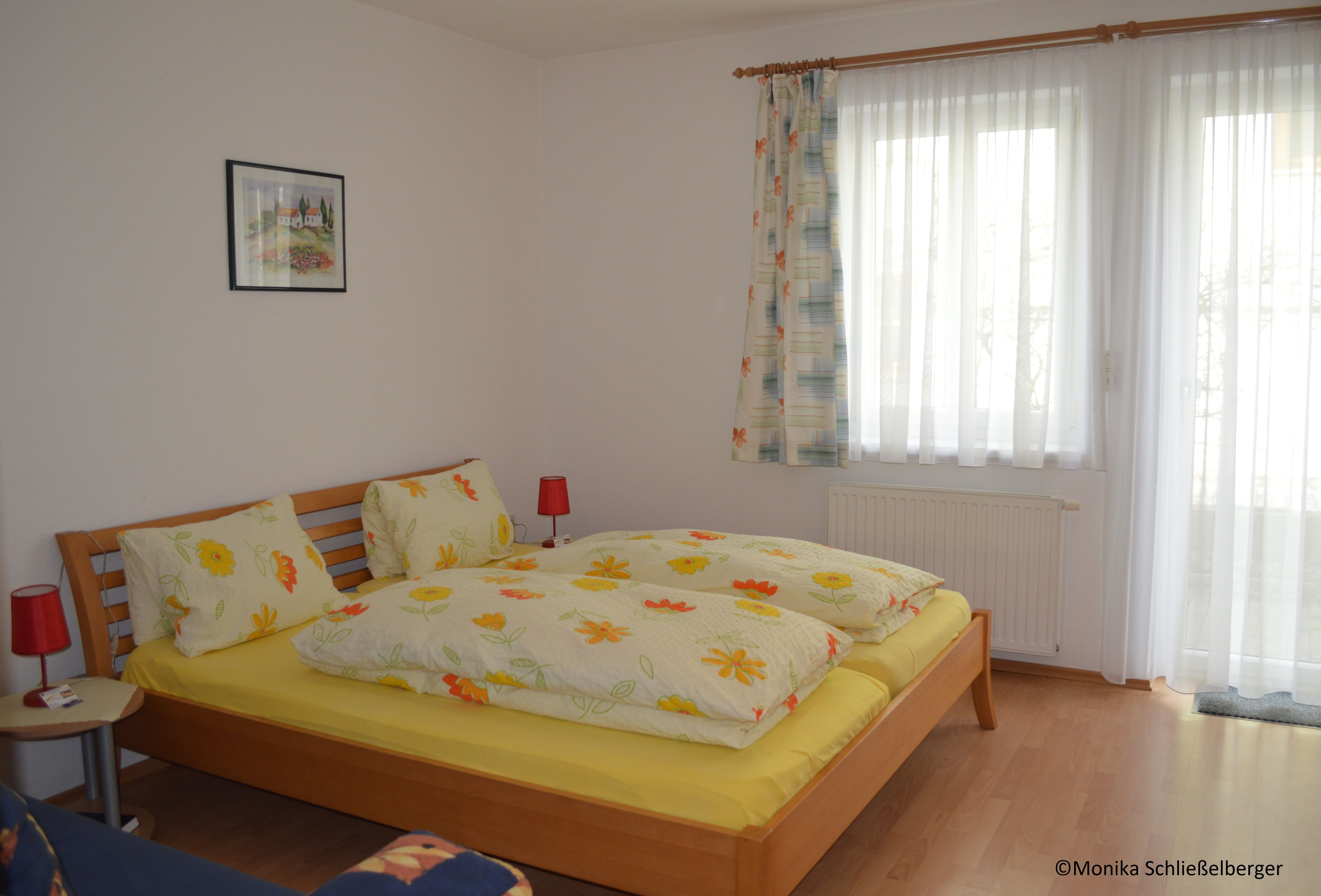 A cozy double room with a wooden bed, colorful bedding and two red bedside lamps. A window with curtains is in the background.