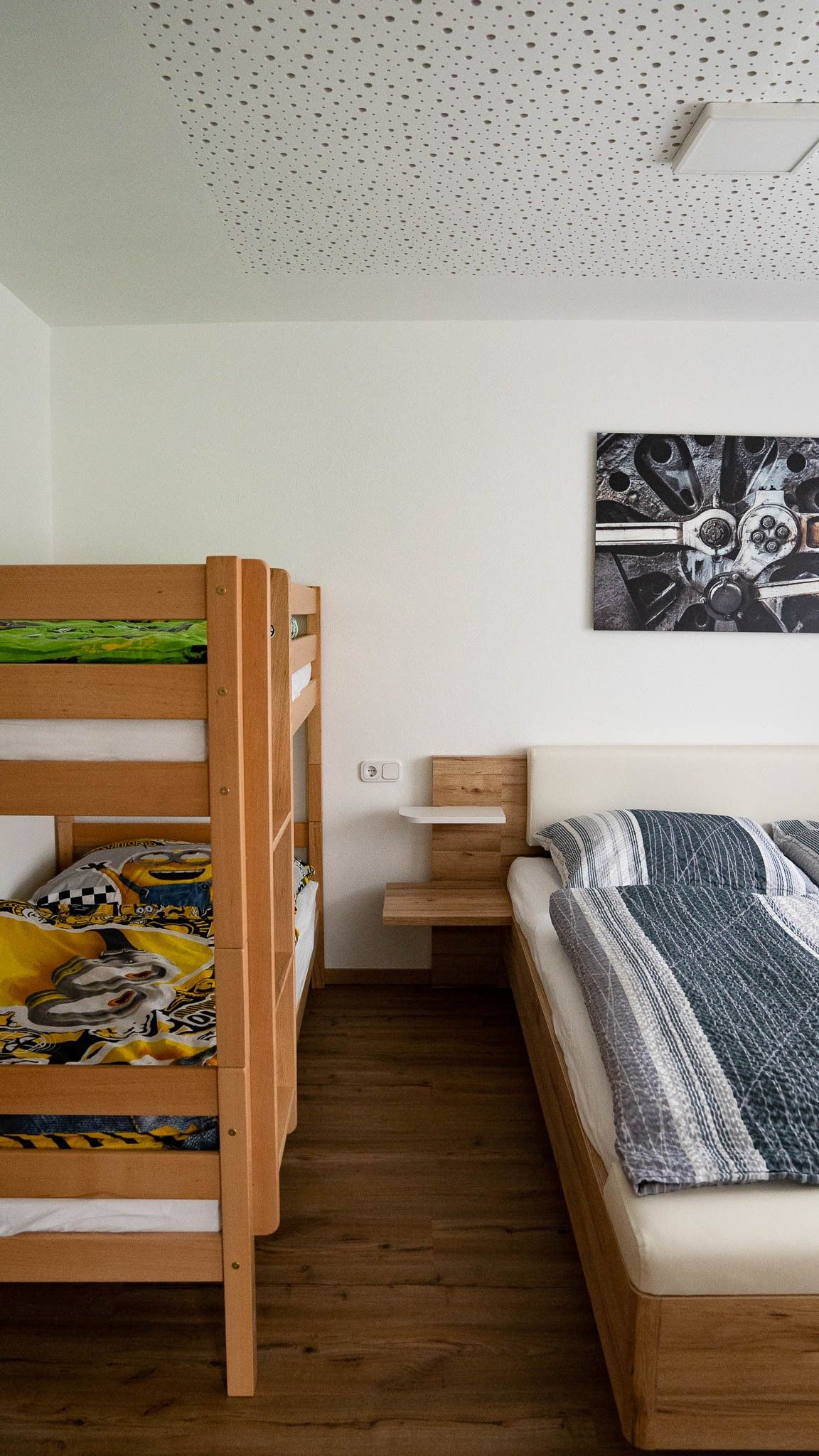 A bedroom with a bunk bed and a double bed made of wood. A picture with a technical motif hangs on the wall.
