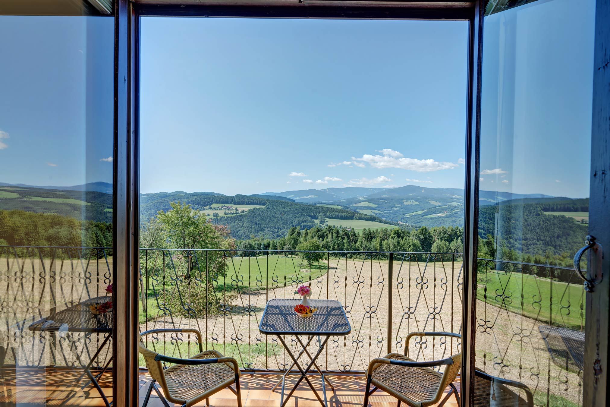 View from a balcony of a hilly landscape with forests and fields, two chairs and a table with flowers in the foreground.