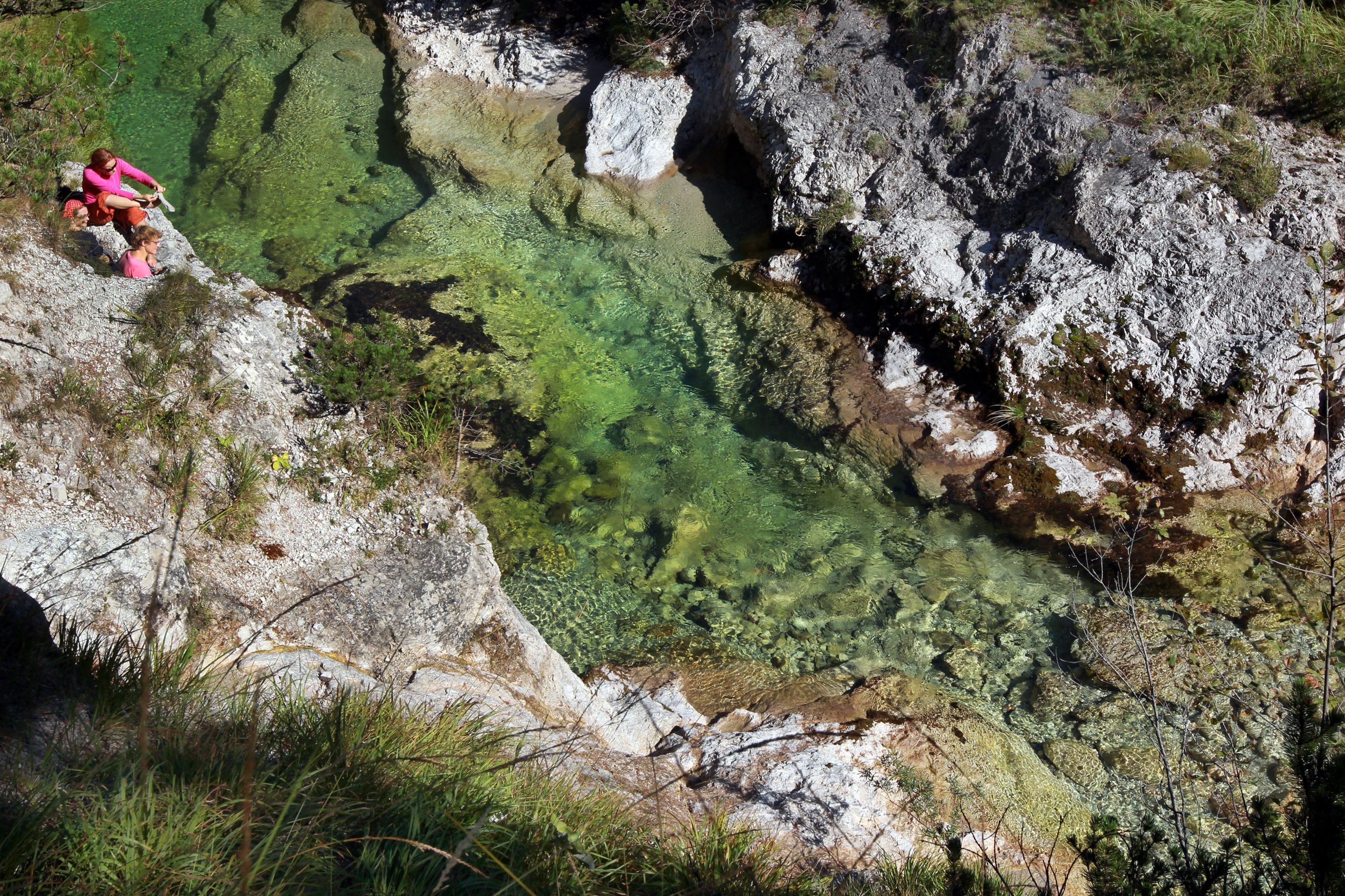 Two people are sitting on a rock next to a clear, green river in a gorge.