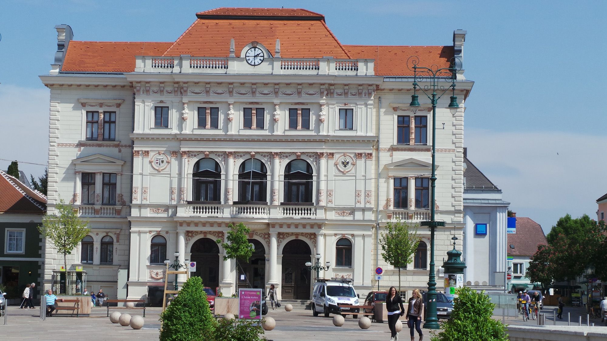 Historic building with clock and decorations, people and cars in the foreground.