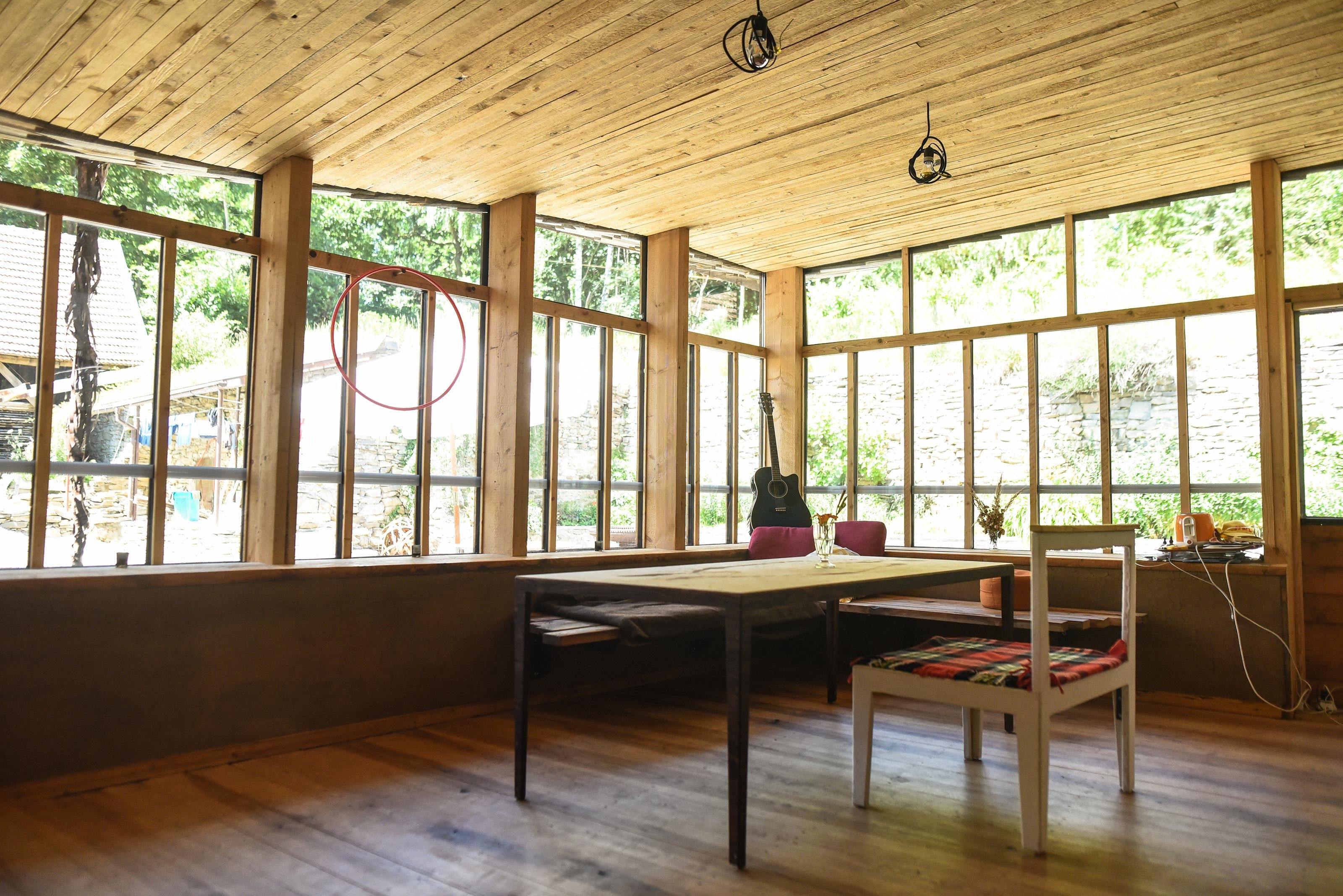A cozy room with wooden floors and a wooden ceiling, large windows, a table, chairs, and a guitar in the corner.