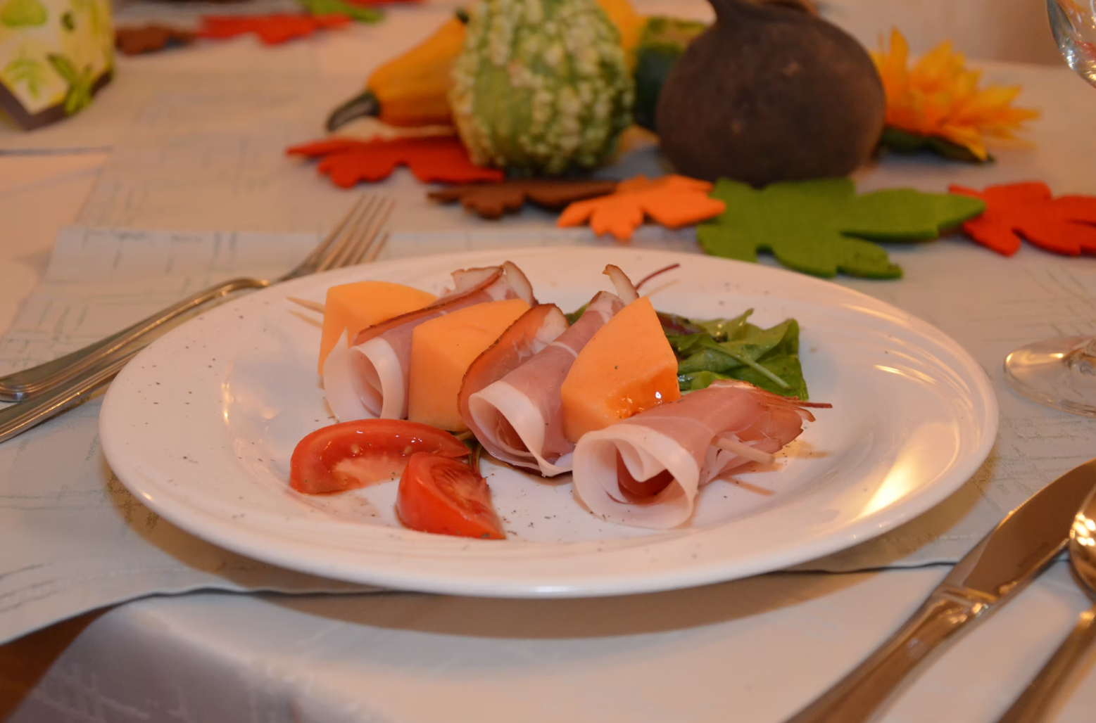 A plate of ham and melon, decorated with tomatoes and salad, on a table with autumnal decorations.
