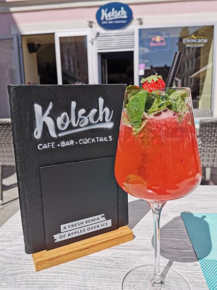 A red cocktail with mint and strawberry in front of a café with the sign 'Kölsch Café Bar Cocktails'.