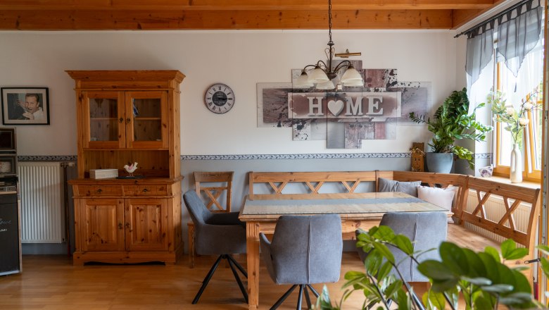 Cozy kitchen with wooden furniture, dining table, grey chairs and plants. A "Home" sign and a clock on the wall.