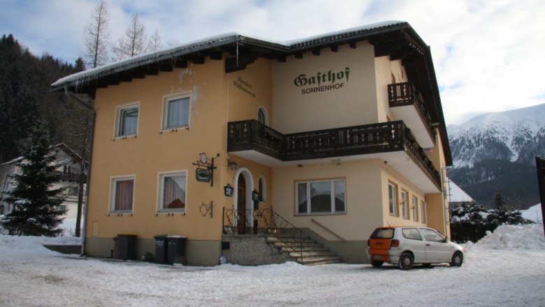 A yellow building with the inscription 'Gasthof Sonnenhof' in the snow, surrounded by mountains.