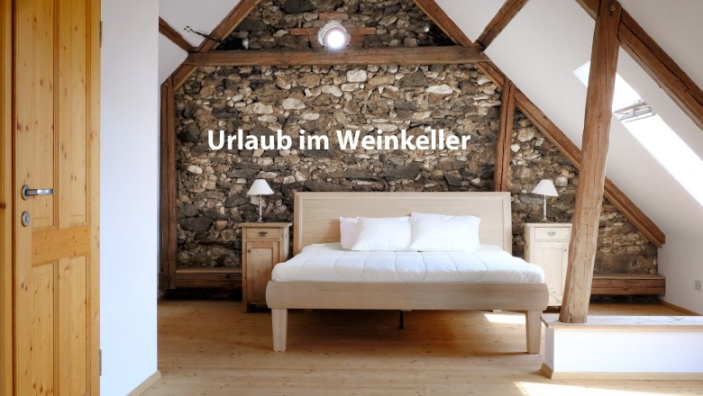 Vacation home "Weinkeller Röschitz", © Weiss und Waldschmidt Bedroom with wooden beams and stone wall, bed in the middle, text 'Vacation in the wine cellar'.