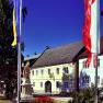 Yellow building with the inscription 'Gasthof Karl Huber', surrounded by flags and a statue.