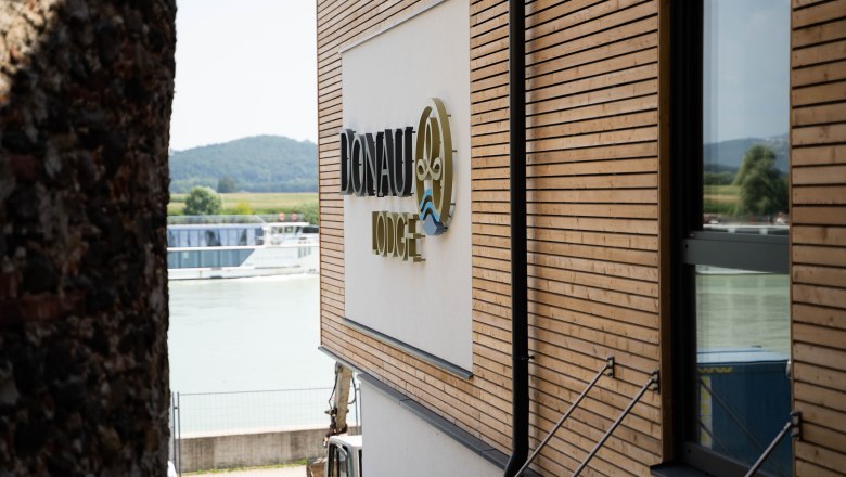 Building with wooden cladding and 'Donau Lodge' lettering, with the Danube and a ship in the background.