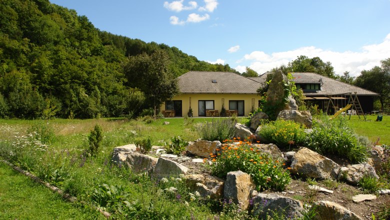 Fischl's guest room, © Archiv Betrieb A yellow house with a garden and rocks in the foreground, surrounded by trees.