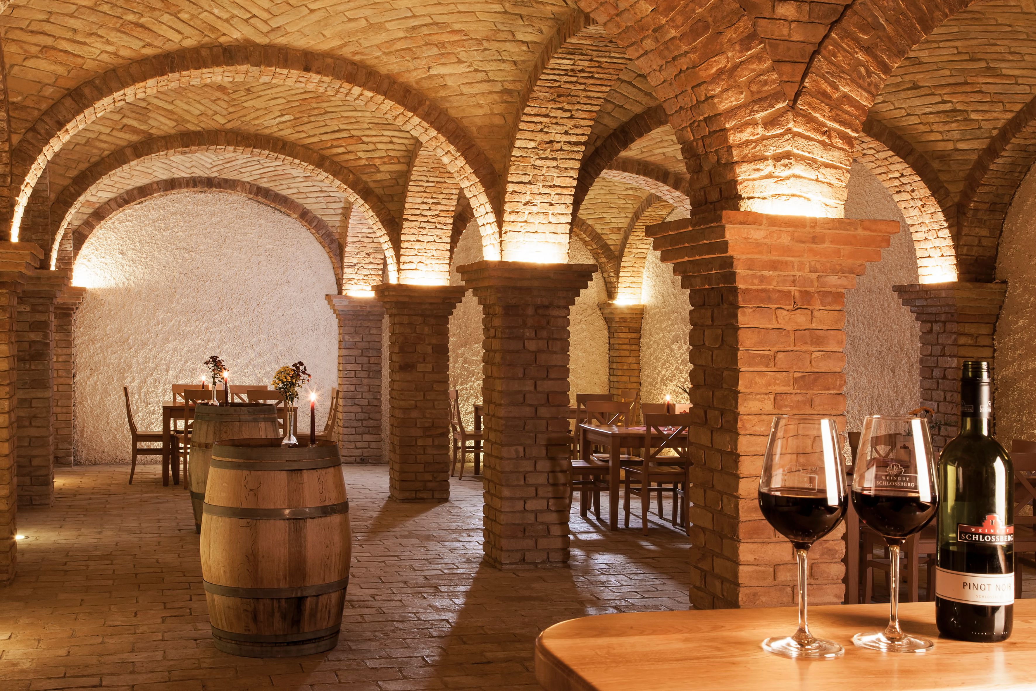 Wine cellar with brick arches, tables and wine barrels, illuminated by candles and lamps.