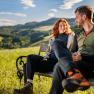 A woman and a man in hiking gear are sitting on a wooden bench. Meadow, green hills and forest in the background.
