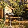 An autumnal garden covered with leaves, a birdhouse in the foreground, a table and chairs in the background, surrounded by trees and a wooden house.
