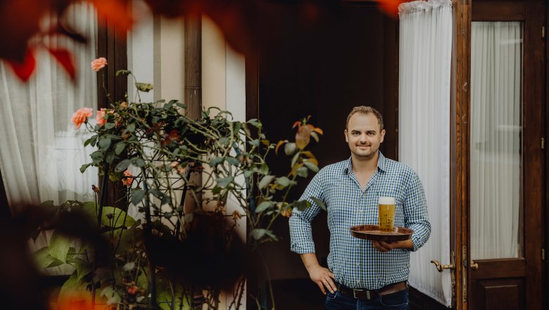 Man with tray and beer in front of a door, surrounded by plants.