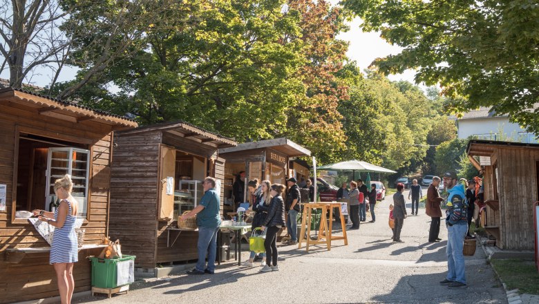 People standing by wooden huts at an outdoor market in sunny weather.