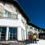 Two people in winter clothing in front of a mountain inn with snow-covered ground and blue sky.