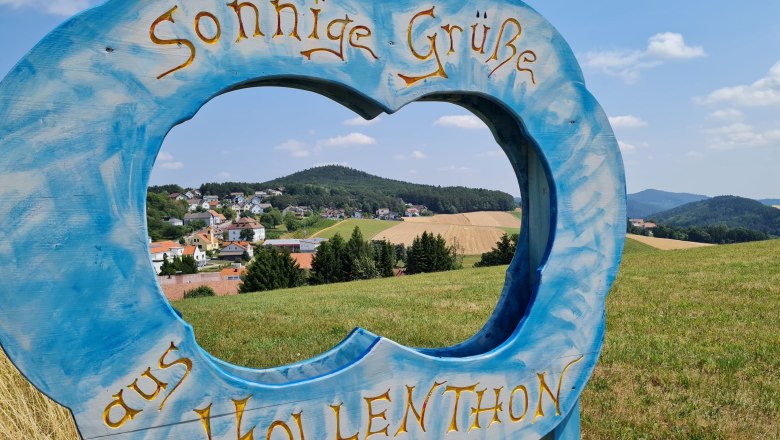Blue wooden frame in the shape of a cloud with the lettering 'Sunny greetings from Hollenthon' in front of a landscape.