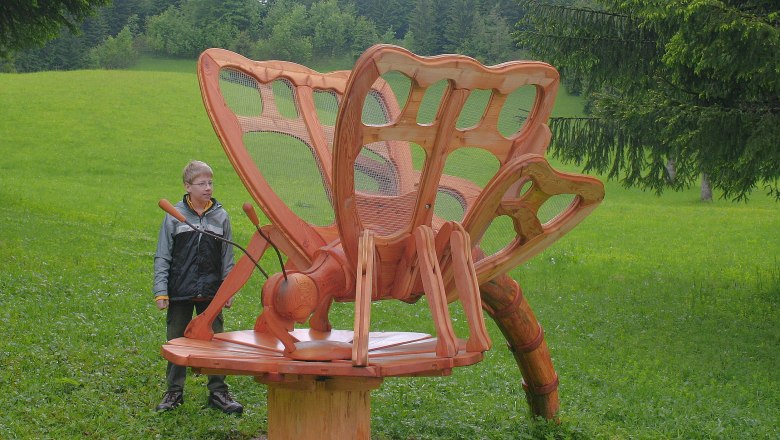 A boy stands next to a large wooden sculpture of a butterfly in a meadow.