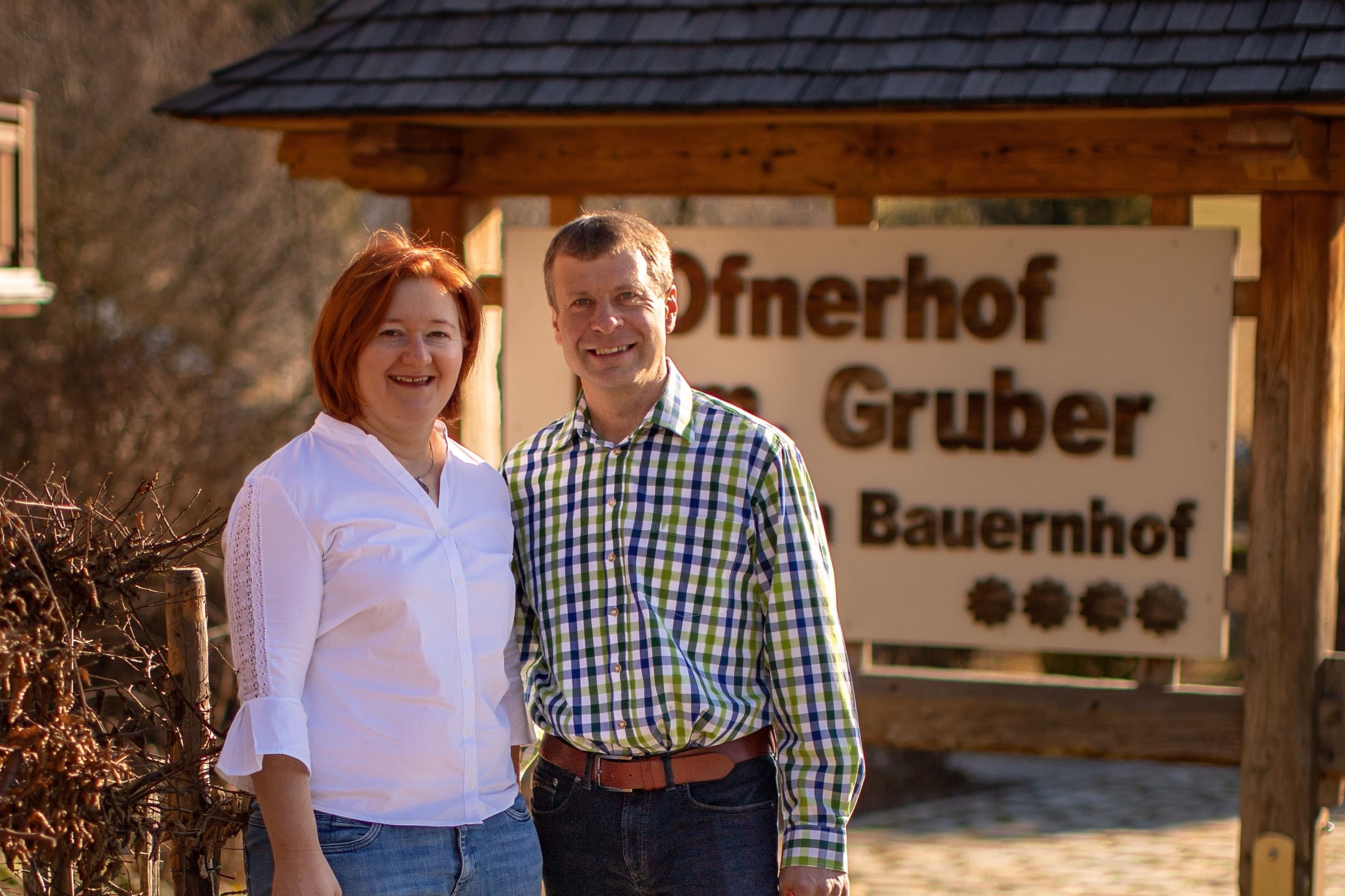 A man and a woman stand smiling in front of a sign that reads 'Ofnerhof Gruber Bauernhof'.