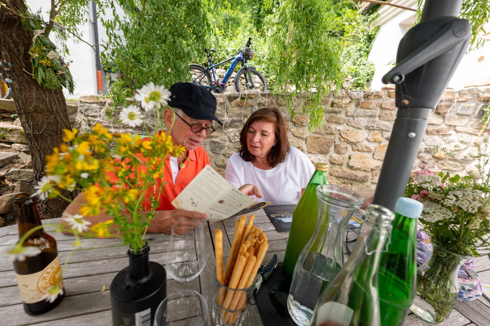 An elderly couple sits at an outdoor table, surrounded by flowers and drinks, looking at a menu.