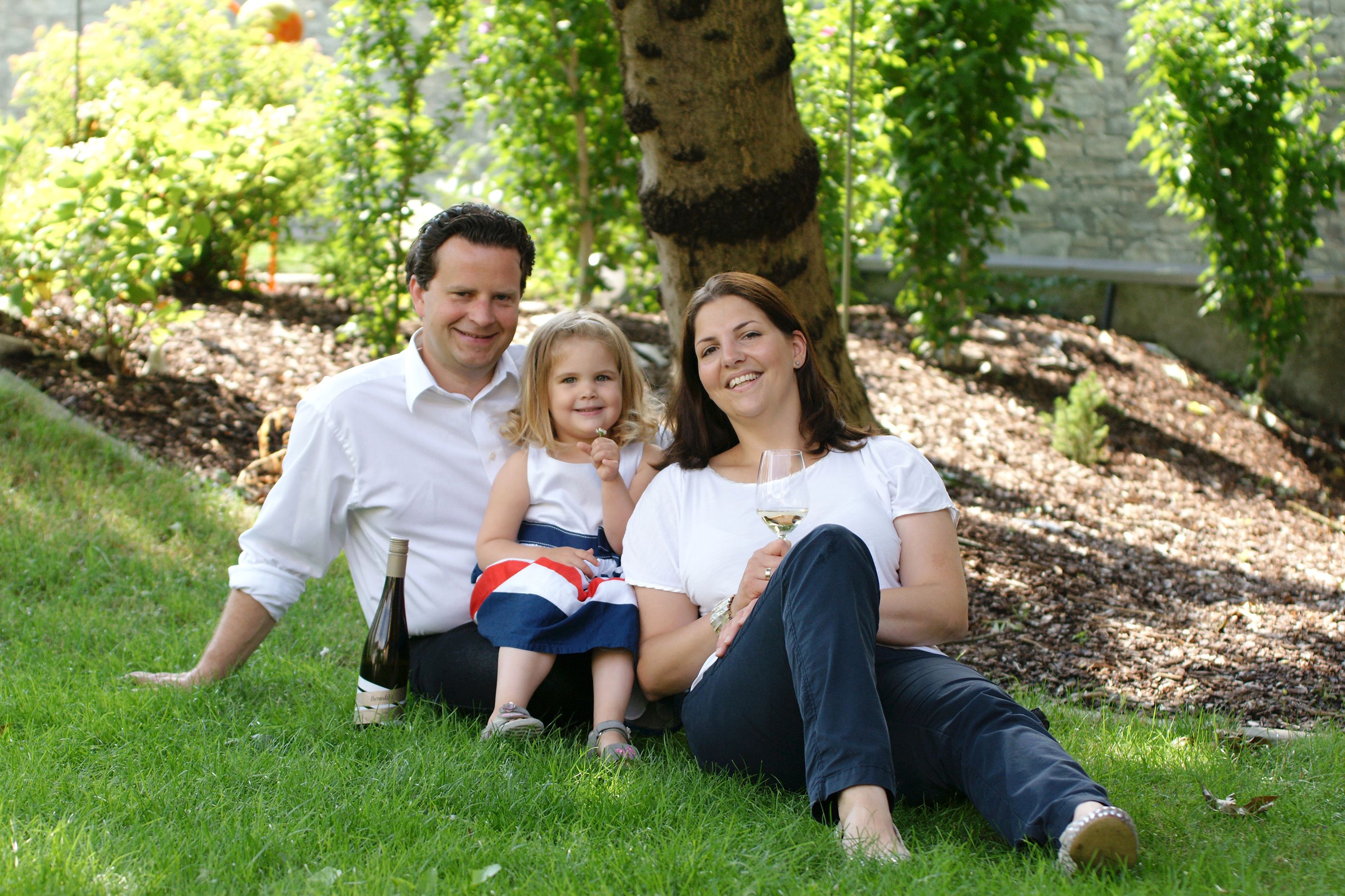 Family sitting in a meadow under a tree with a bottle of wine and a glass.