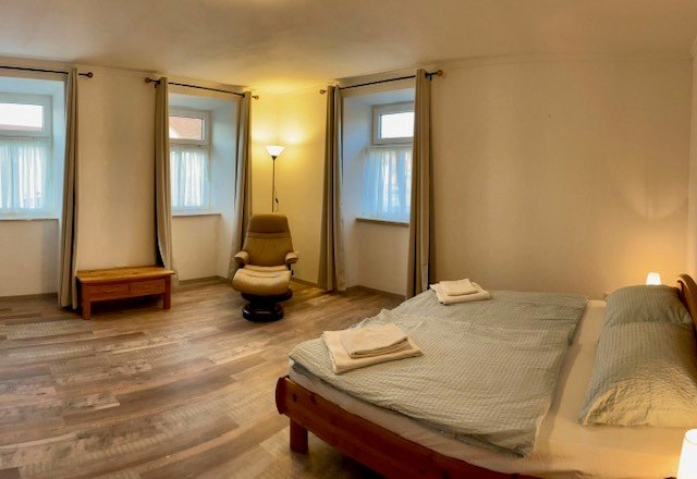 A simple bedroom with double bed, armchair, bedside table and three windows with curtains.