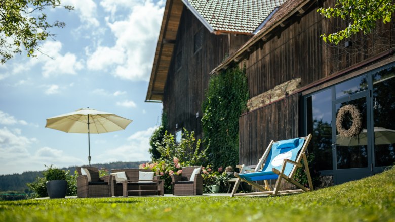A farmhouse with a wooden façade, garden furniture and parasol on a meadow in sunny weather.