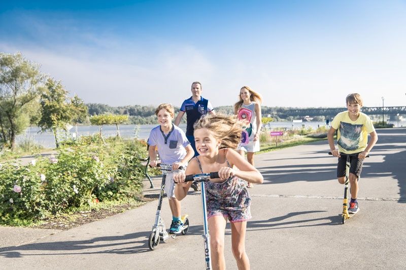 Children ride scooters on a path along a river, accompanied by two adults in the background.