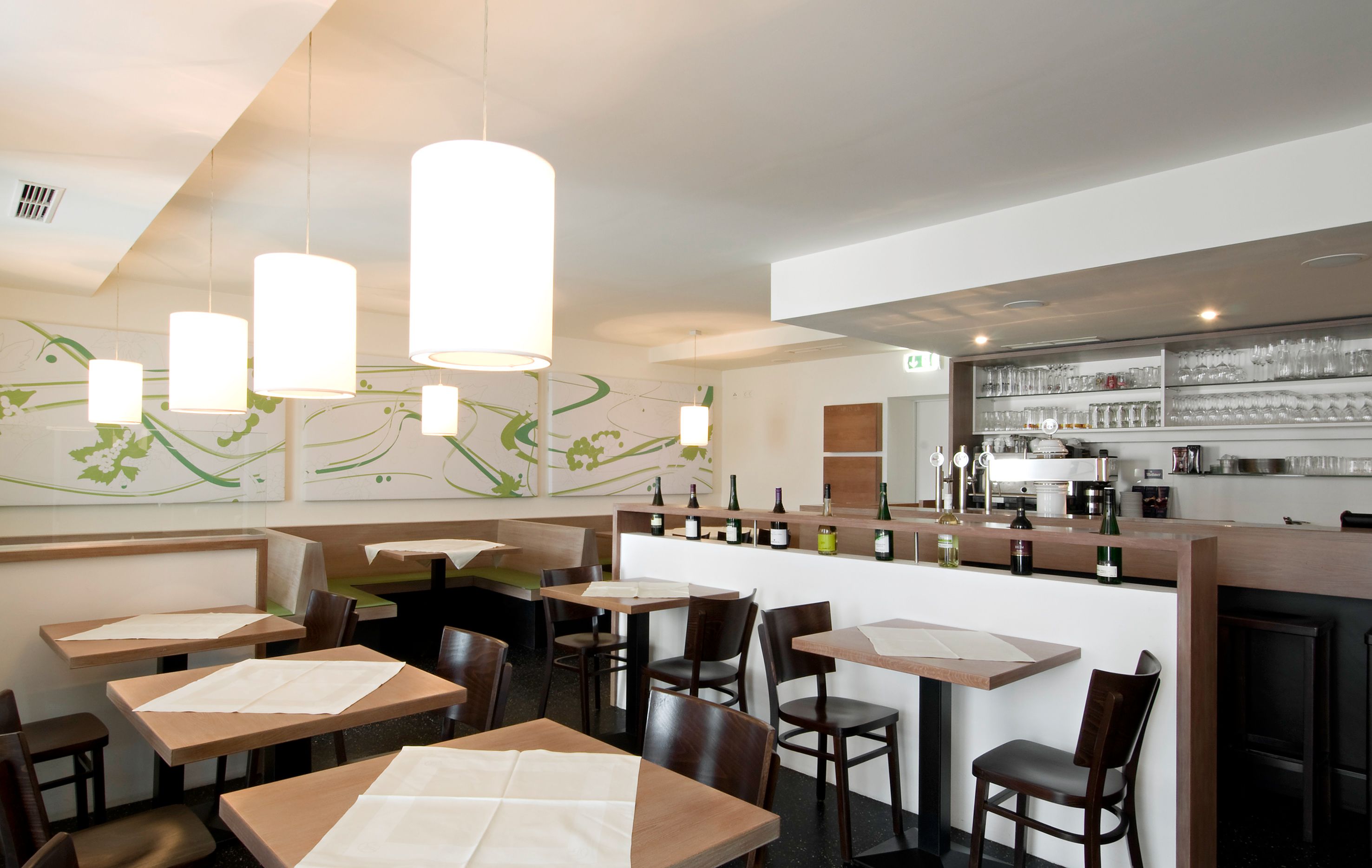 Interior view of a modern restaurant with wooden tables, chairs and a bar with bottles and glasses.
