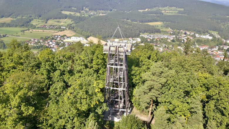 Observation tower surrounded by trees with a view of a village and wooded hills in the background.