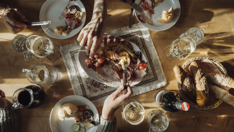 A table with a Heurigen snack consisting of bread, cold cuts, cheese and wine, surrounded by several hands.