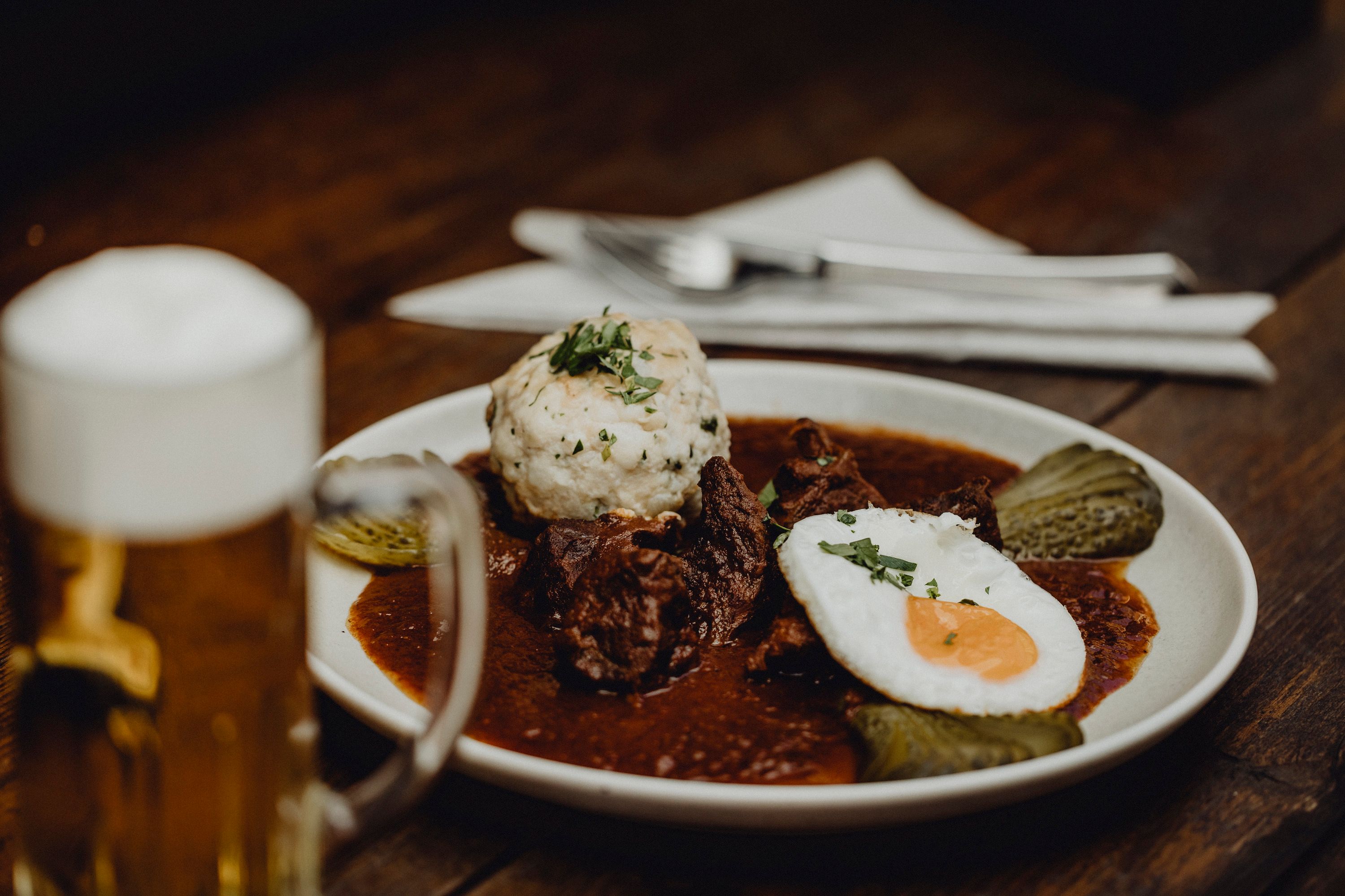 A plate with goulash, dumplings, fried egg and gherkins, next to it a beer mug on a wooden table.