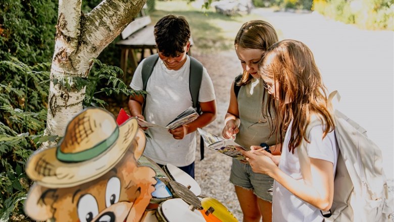 Three children with rucksacks reading cards next to a bear figure outdoors.